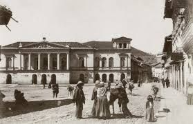 Imagen antigua en blanco y negro que muestra una plaza pública con personas vestidas con ropas tradicionales, representando la vida cotidiana en una ciudad histórica.