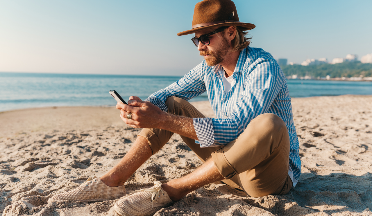 Hombre en la playa utilizando su teléfono móvil para la planificación de presupuesto vacacional mediante herramientas digitales.