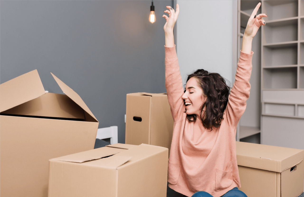 Mujer joven sonriente levantando los brazos rodeada de cajas de mudanza, representando la independencia y la importancia de ahorrar para independizarte."