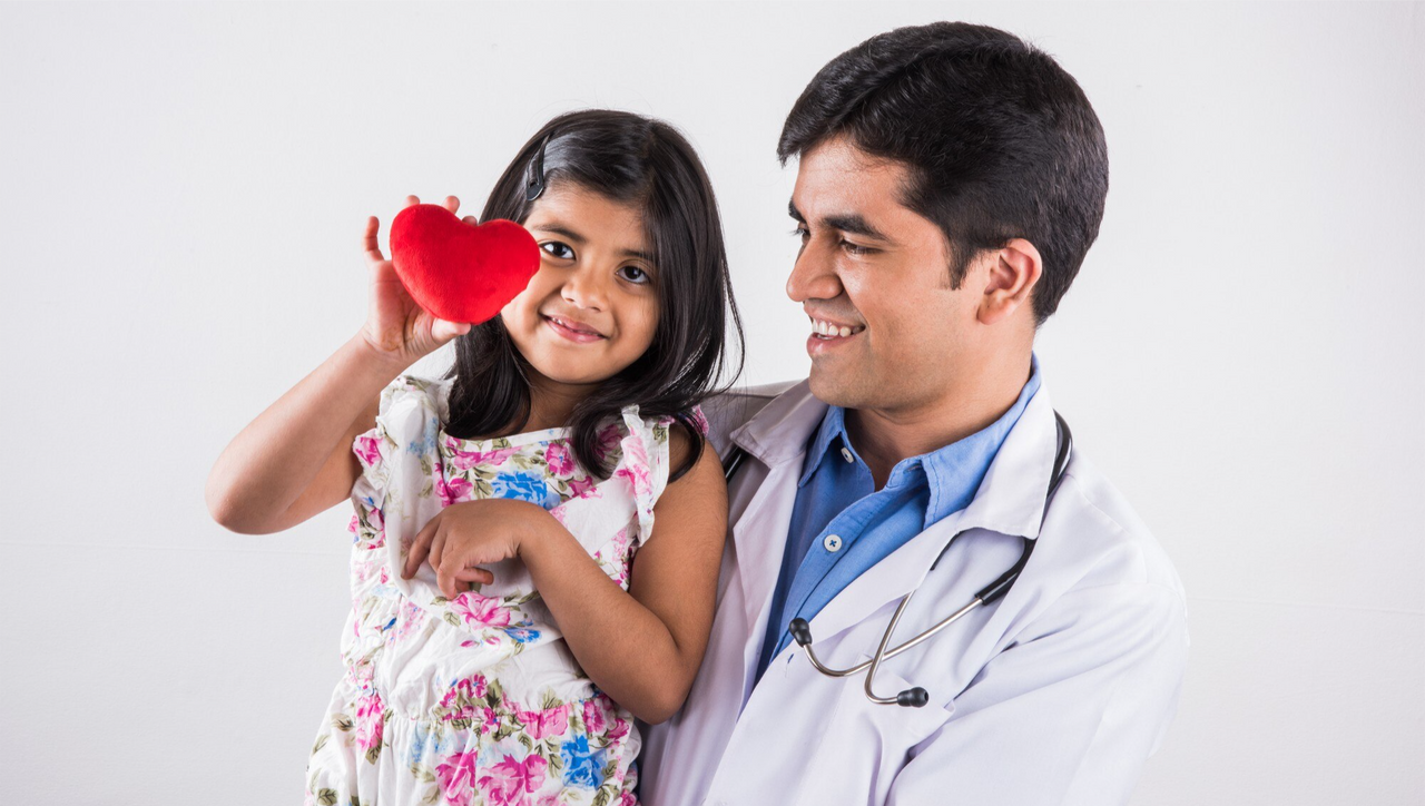 Un médico sosteniendo a una niña pequeña que sonríe mientras sostiene un corazón rojo, representando cuidado, amor y protección familiar."