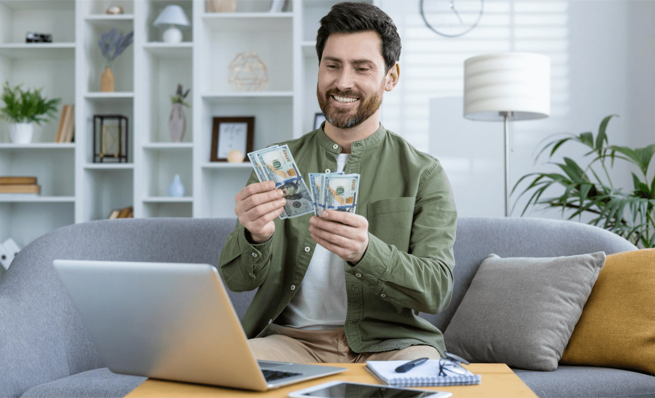 Hombre sonriente contando billetes de dólar frente a su laptop en casa, simbolizando éxito financiero y buen manejo de finanzas personales