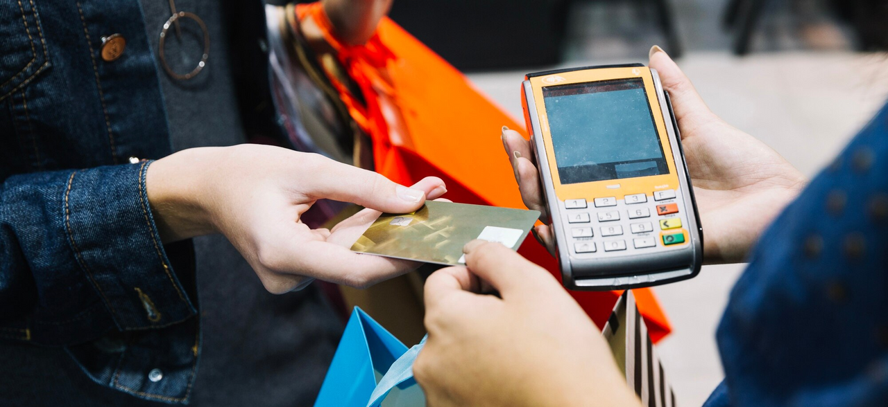 Persona realizando un pago con tarjeta de crédito en un datáfono durante una jornada de compras, con bolsas y artículos alrededor.