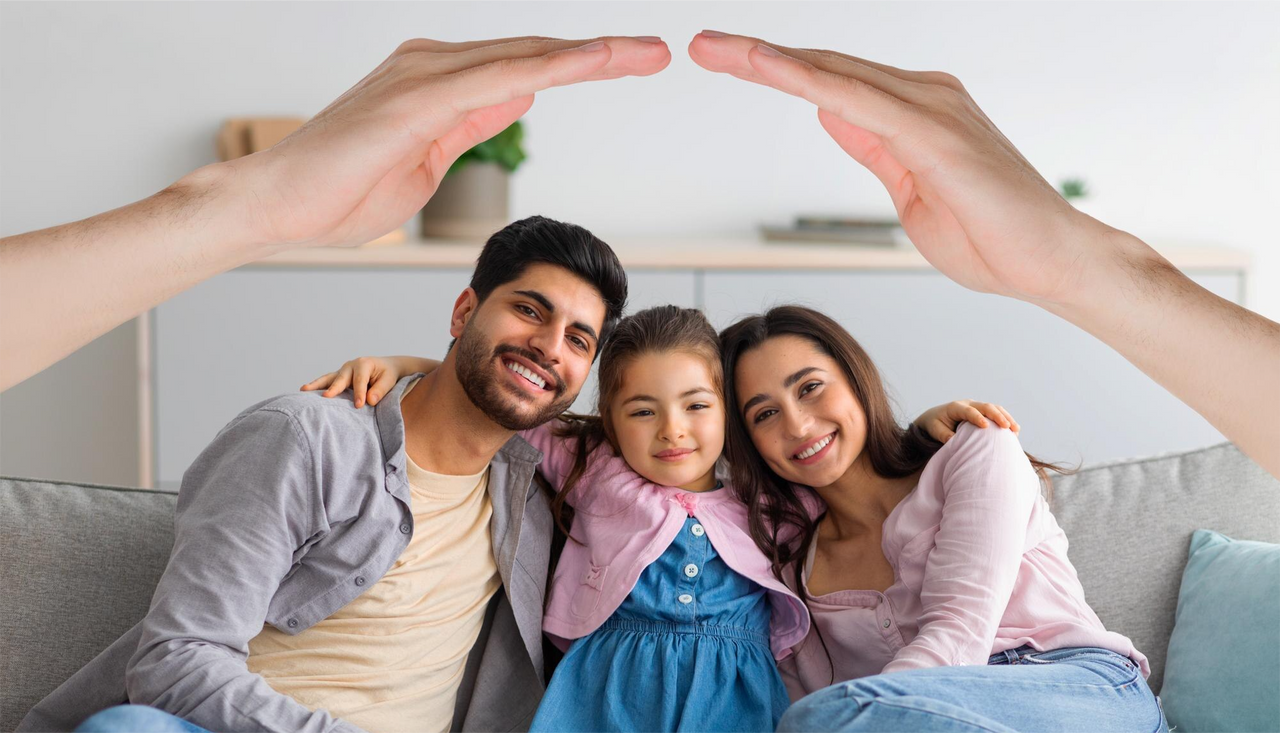 Familia sonriente compuesta por padre, madre e hija abrazados en un sofá, protegidos simbólicamente por unas manos que forman un techo.