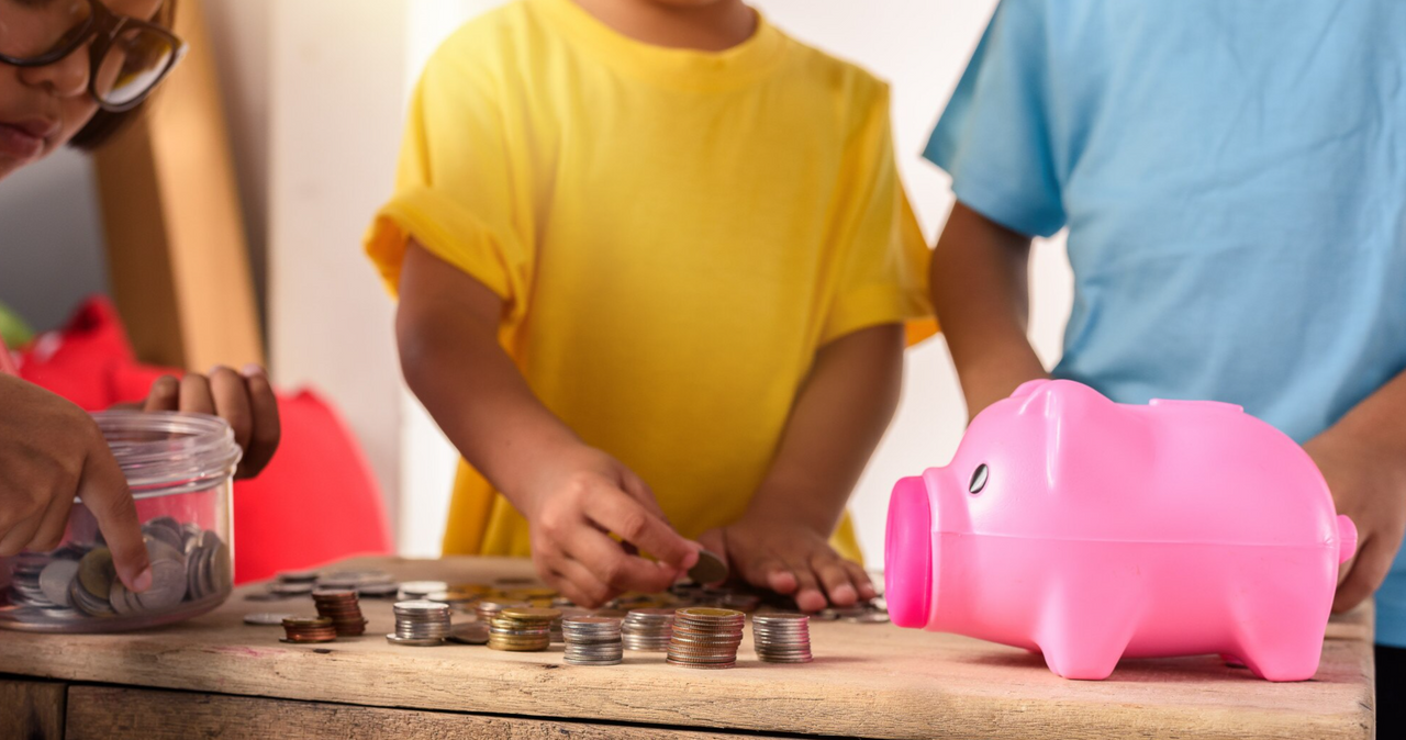 Tres niños participando en una actividad de educación financiera, contando y organizando monedas sobre una mesa de madera junto a una alcancía rosada en forma de cerdito.