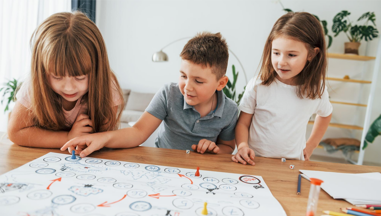 Tres niños —dos niñas y un niño— se encuentran alrededor de una mesa de madera, concentrados mientras juegan un juego de mesa con fichas, dados y casillas numeradas. 