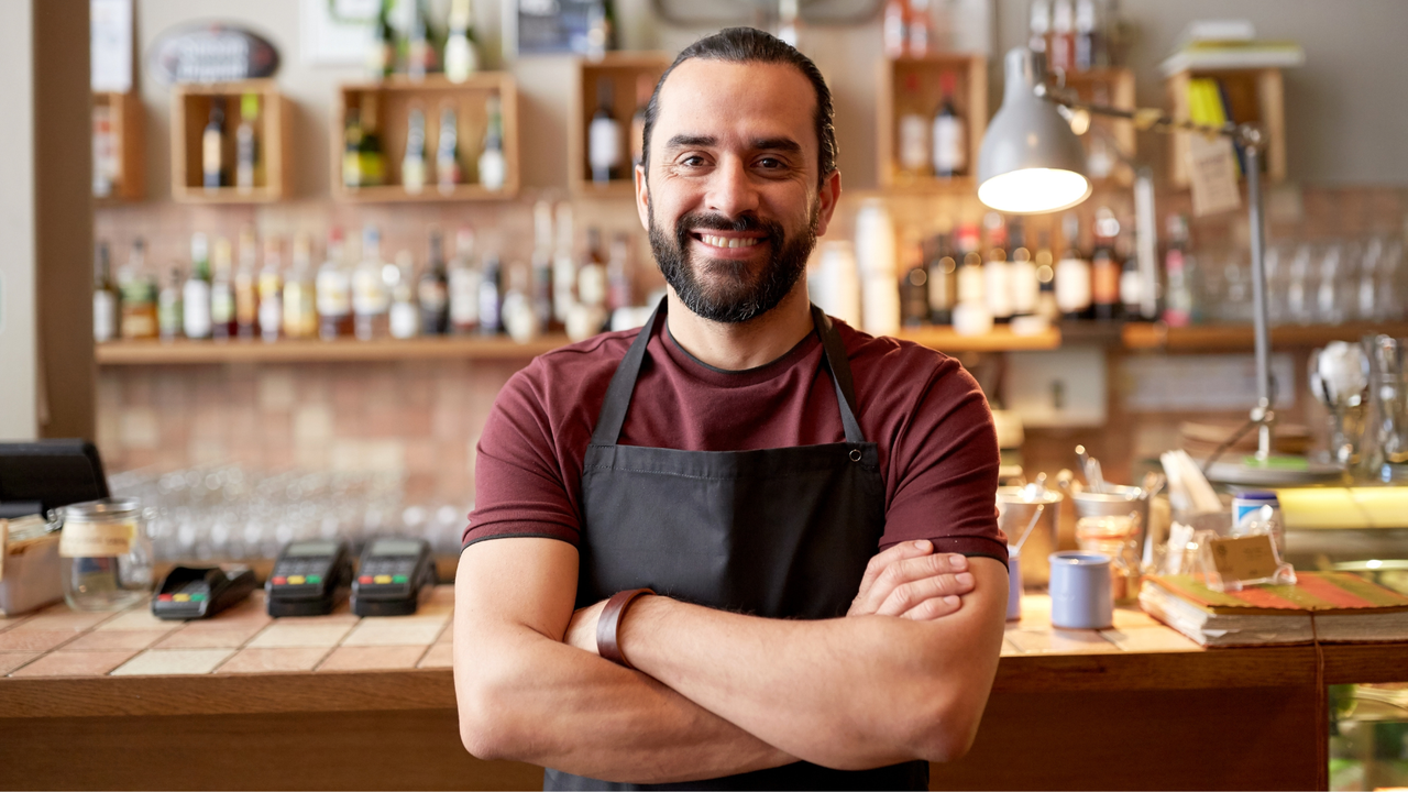 Emprendedor sonriente con mandil negro, de brazos cruzados, dentro de su cafetería con barra y estantes de bebidas al fondo.